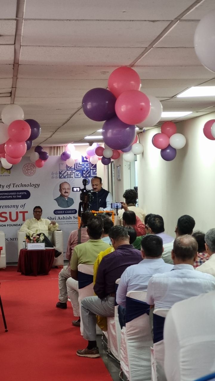 A full room of guests seated under pink and purple balloons at the AIC NSUT inauguration ceremony, with the chief guest speaking at the front and a camera recording