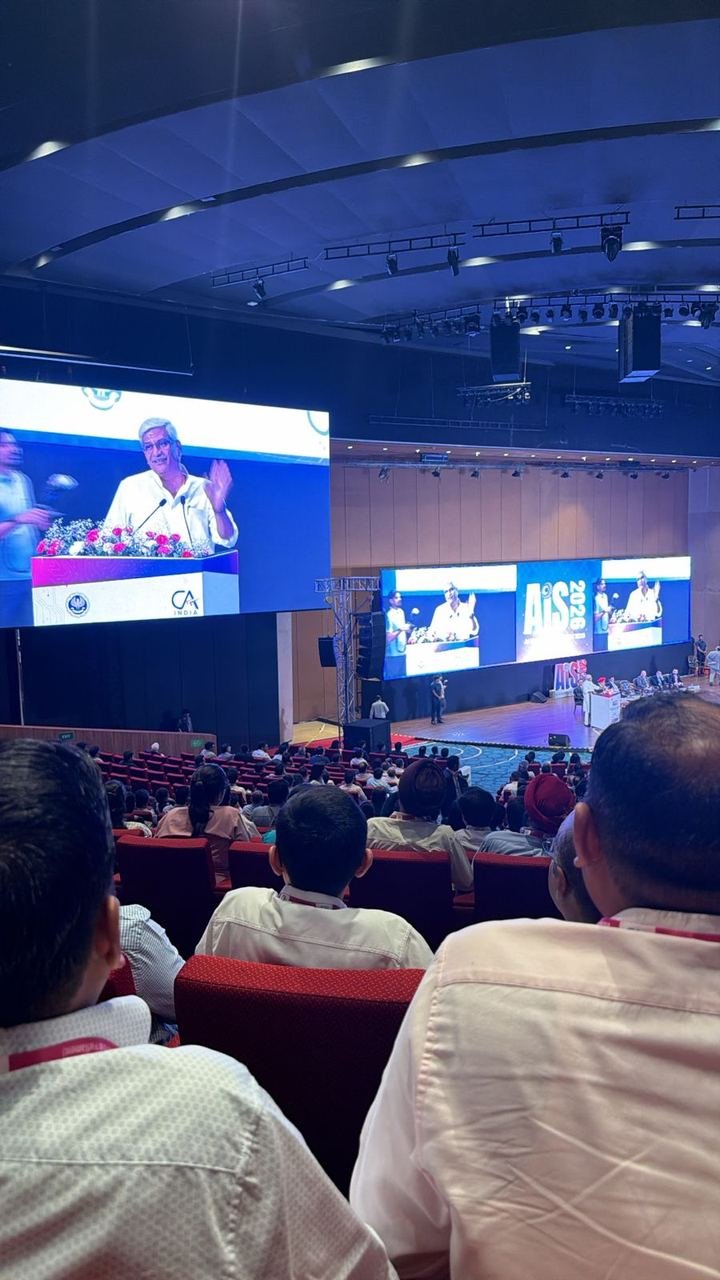 Delegates with lanyards filling the red seats of the AIS 2026 main hall at Bharat Mandapam, watching a speaker on the large screens beside the stage