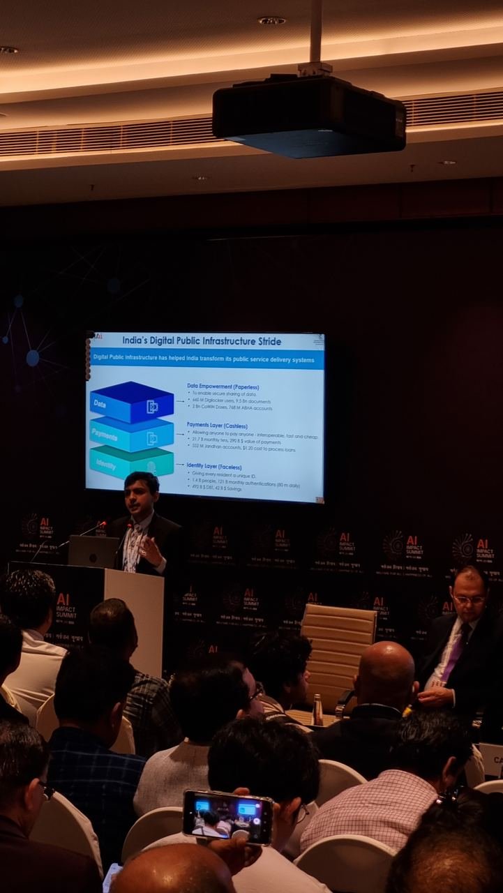 A speaker at a summit session presenting a slide titled India's Digital Public Infrastructure Stride, showing identity, payments and data layers, to a full room