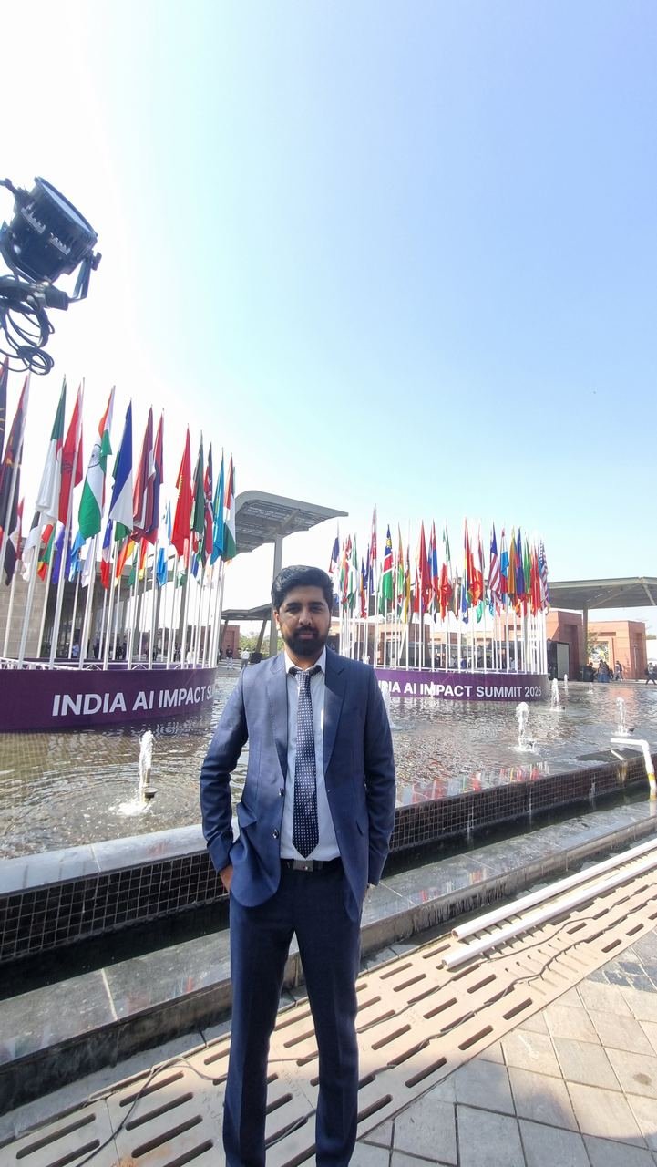 A Zoffer team member in a blue suit standing by the fountain in front of the ring of national flags and the India AI Impact Summit 2026 sign at Bharat Mandapam