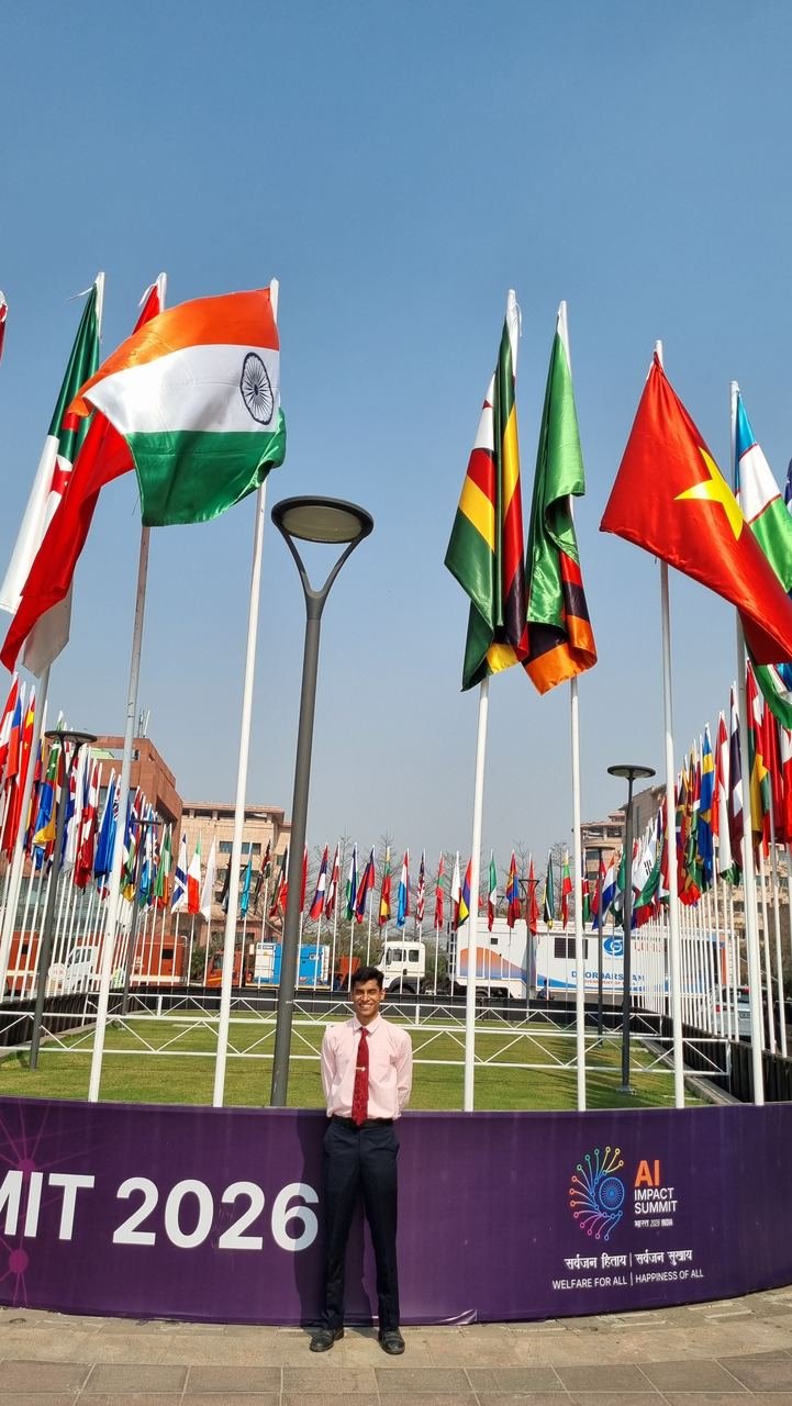 A Zoffer team member in a pink shirt and red tie standing at the AI Impact Summit 2026 banner, beneath a circle of national flags including India's